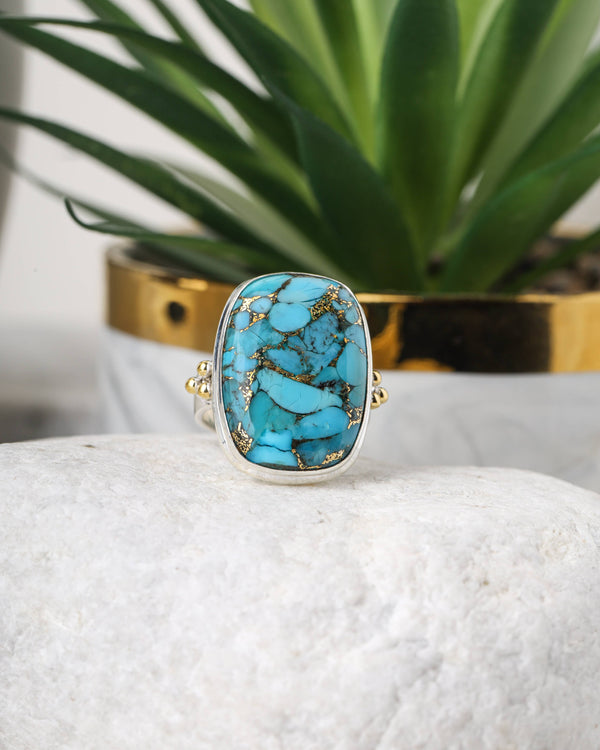 Turquoise ring with gold accents on a white surface with a plant in the background