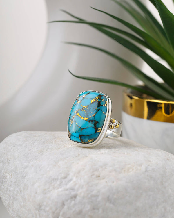 Turquoise ring with gold accents on a white stone surface with a plant in the background