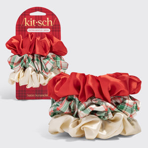 A set of four Kit Sch satin scrunchies displayed on a light gray background, with the retail packaging card standing behind them. The scrunchies include a large red satin scrunchie, a green and red plaid satin scrunchie, a cream satin scrunchie, and a matching plaid and satin combination.